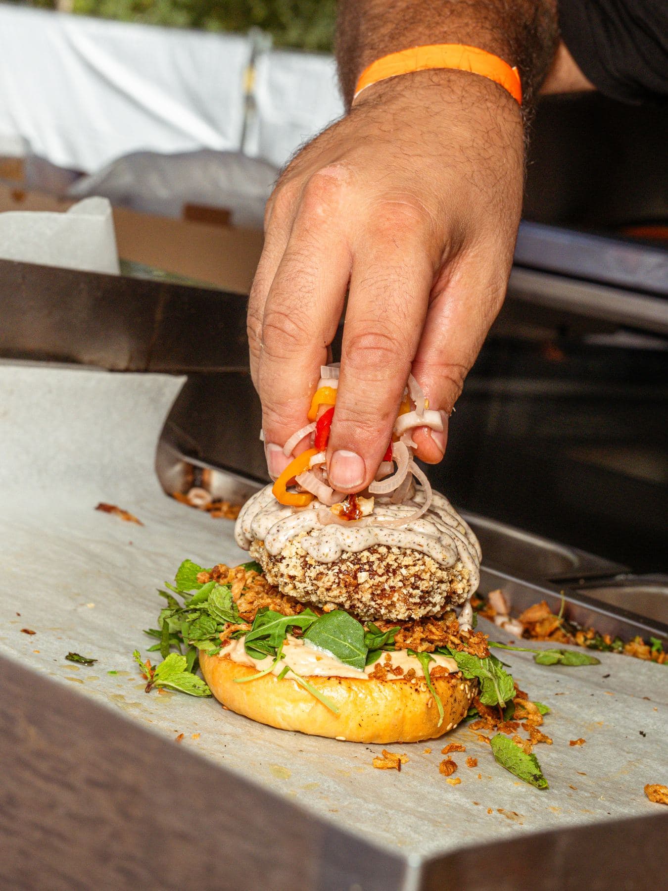 Een hand legt de laatste ingrediënten op een burger, op het RVS-werkblad van een foodtrailer